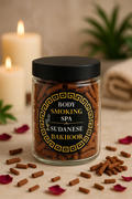 A clear glass jar filled with brown smoking spa sticks, topped with a black lid and labeled 'BODY SMOKING SPA SUDANESE BAKHOOR,' set on a beige surface with scattered sticks, rose petals, candles, and a soft spa-like background.